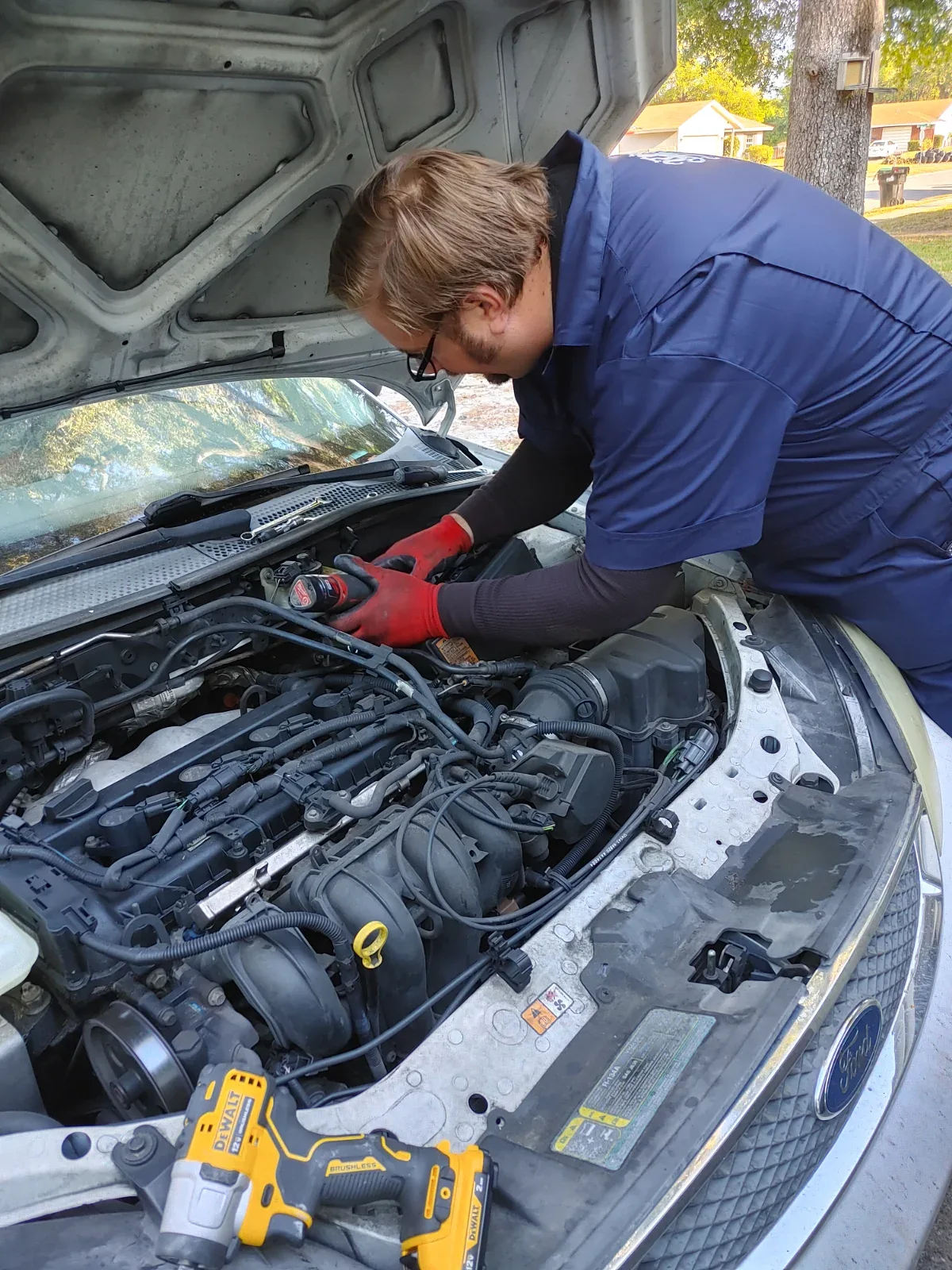 A person in a blue uniform and red gloves works on a car engine with the hood open.