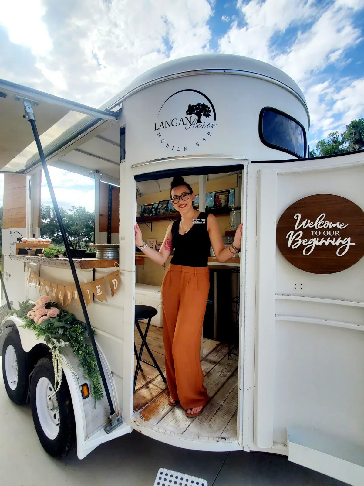 A smiling woman stands in the open doorway of a white mobile bar trailer with "LANGAN Acres MOBILE BAR" on its side.