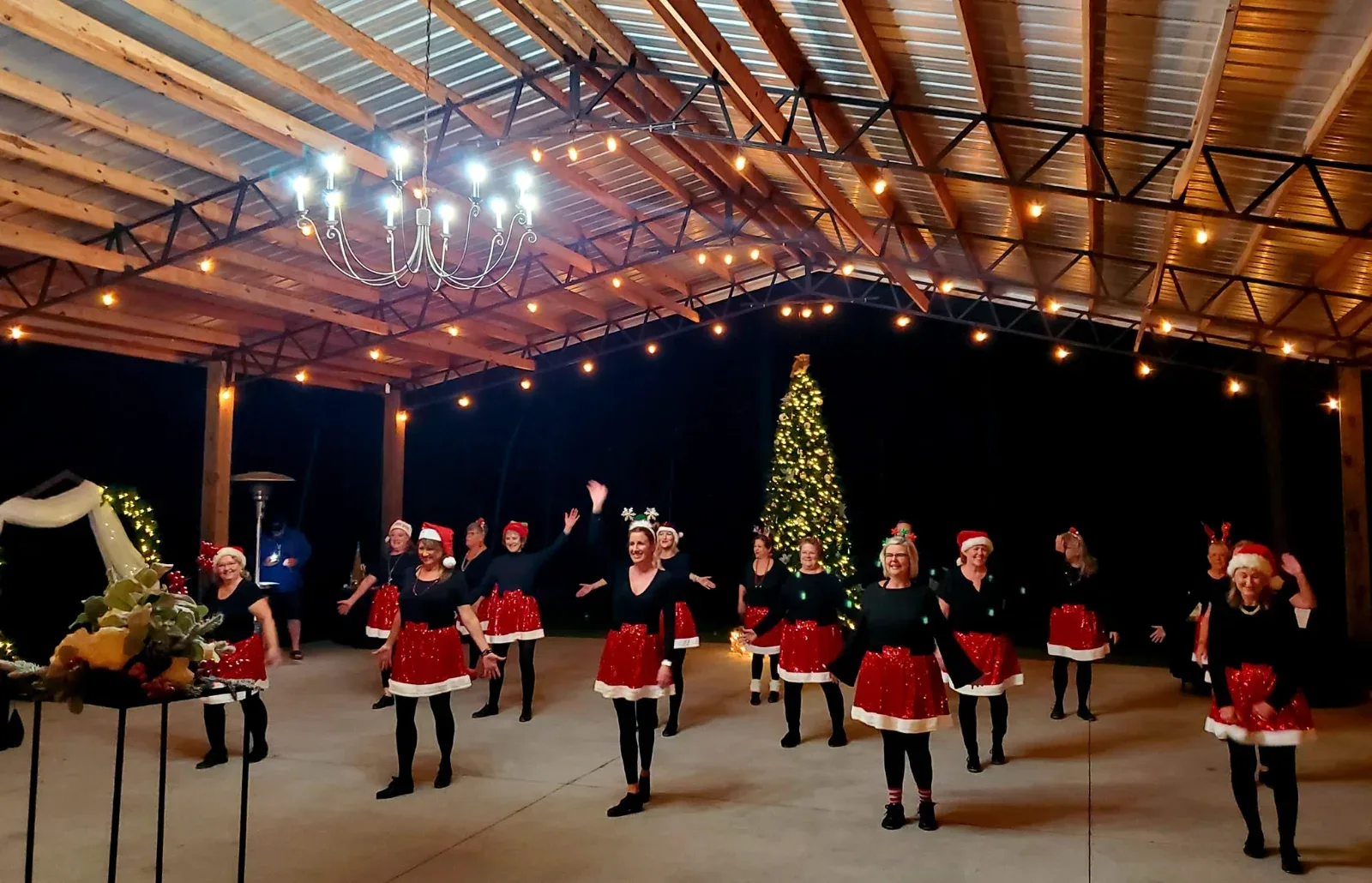 A group of women in red and black holiday outfits dancing under a lit pavilion with a Christmas tree.