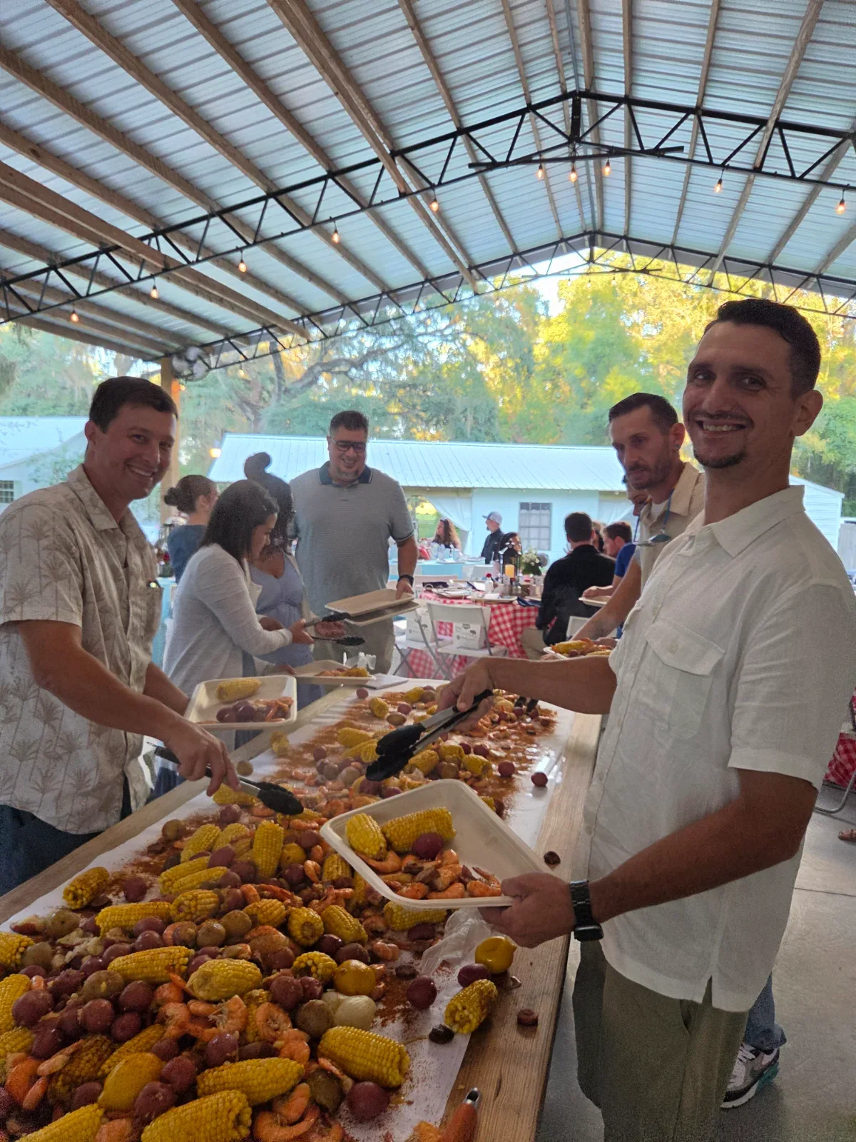 People serving themselves from a long table covered with corn, potatoes, and shrimp.