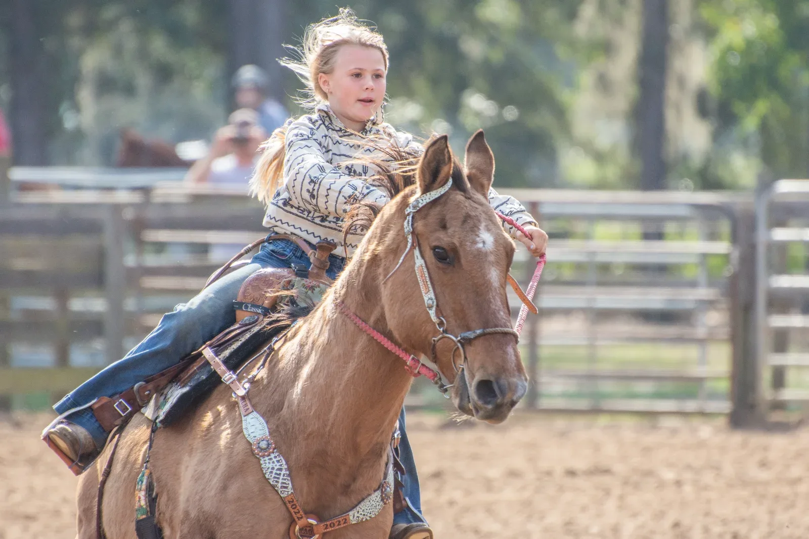 A young person in a patterned shirt and jeans rides a brown horse with a pink bridle.