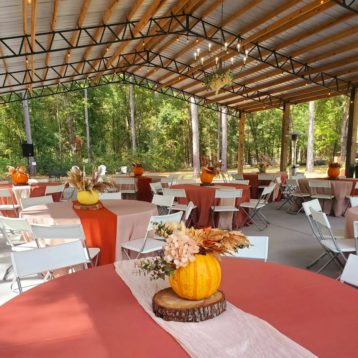 An outdoor pavilion with a metal roof, string lights, and round tables set with fall-themed centerpieces.