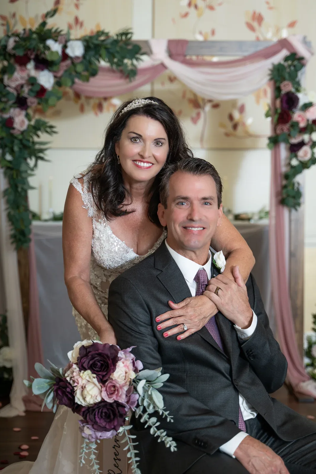 A smiling man and woman in formal attire, with the woman holding a bouquet of purple and pink flowers.