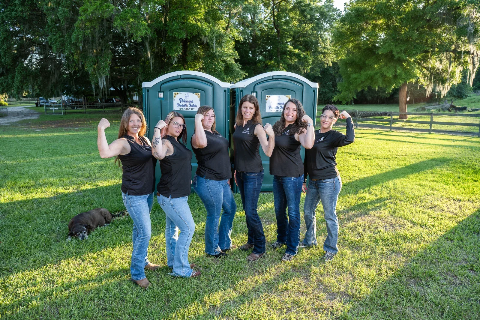Six women in black shirts and jeans flexing their biceps in front of two green portable toilets.