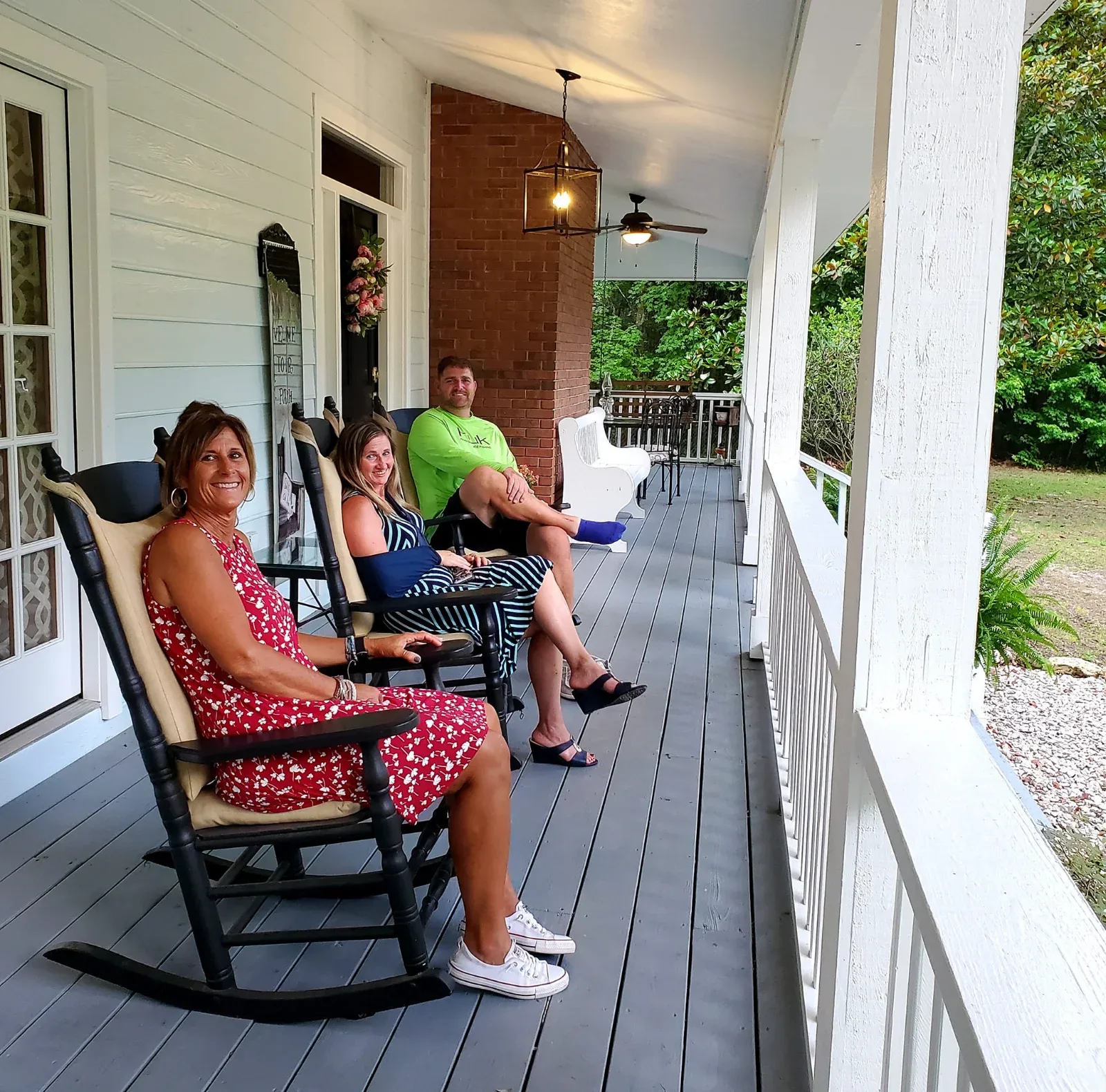Three people sit in rocking chairs on a porch with white railings and a brick wall.