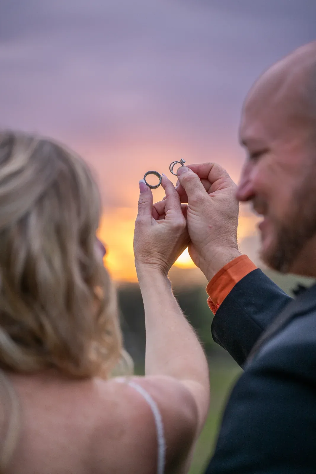 Two people hold up rings against a sunset sky.