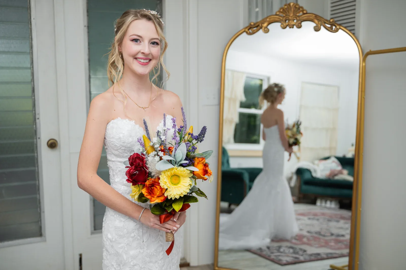 A smiling woman in a white dress holds a colorful bouquet, her reflection visible in a gold mirror.