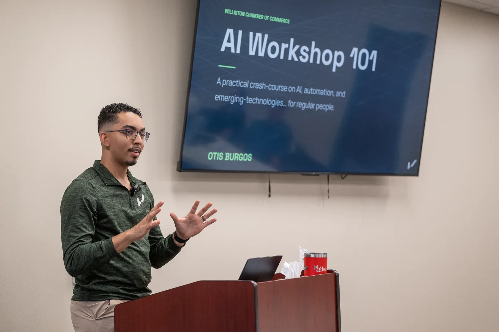 A man speaks at a podium in front of a screen displaying "AI Workshop 101" and "OTIS BURGOS".