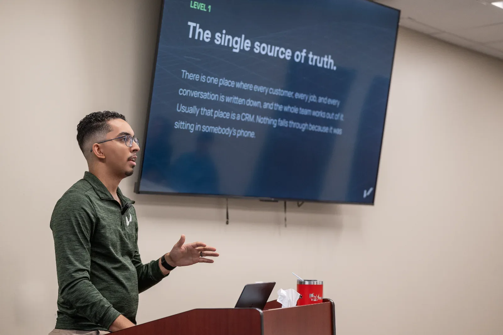 A man in glasses speaks at a podium with a laptop, tissues, and a red mug, facing a screen displaying "The single source of truth.
