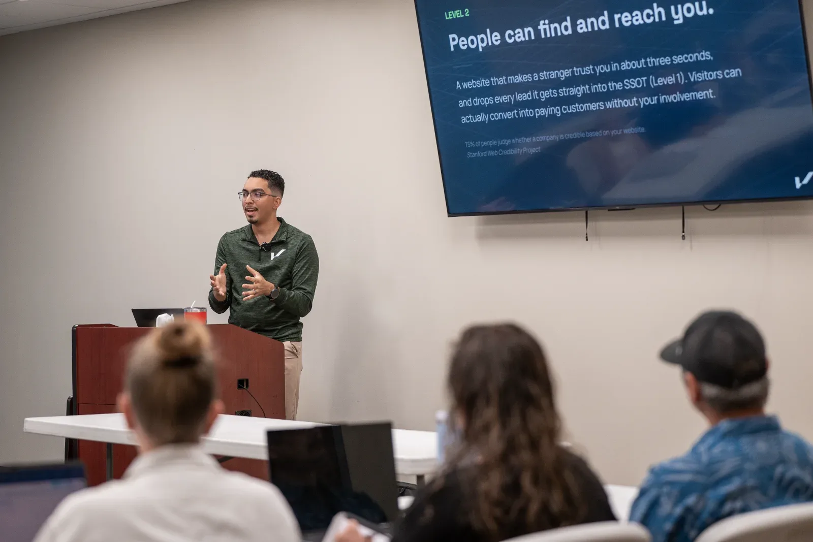 A man speaks at a podium to an audience, with a screen displaying "People can find and reach you.