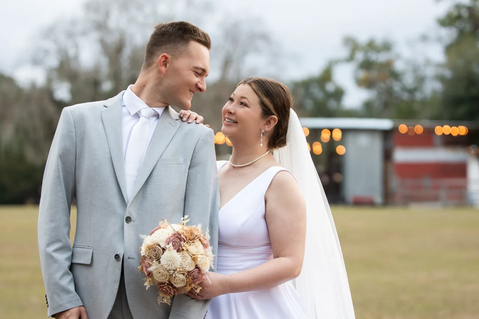 A man in a gray suit and a woman in a white dress with a bouquet, smiling at each other.