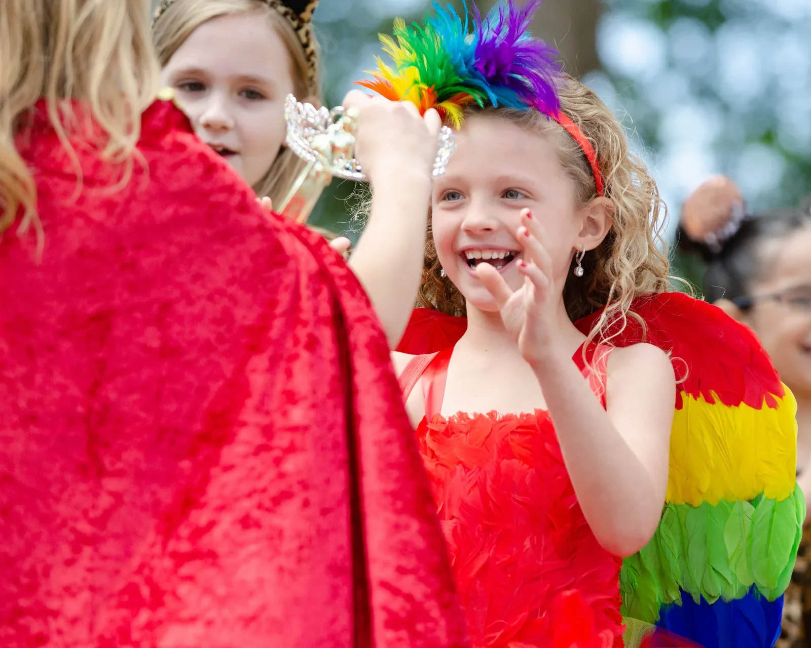 A person in a red cape places a crown on a smiling girl with a rainbow feather headband.