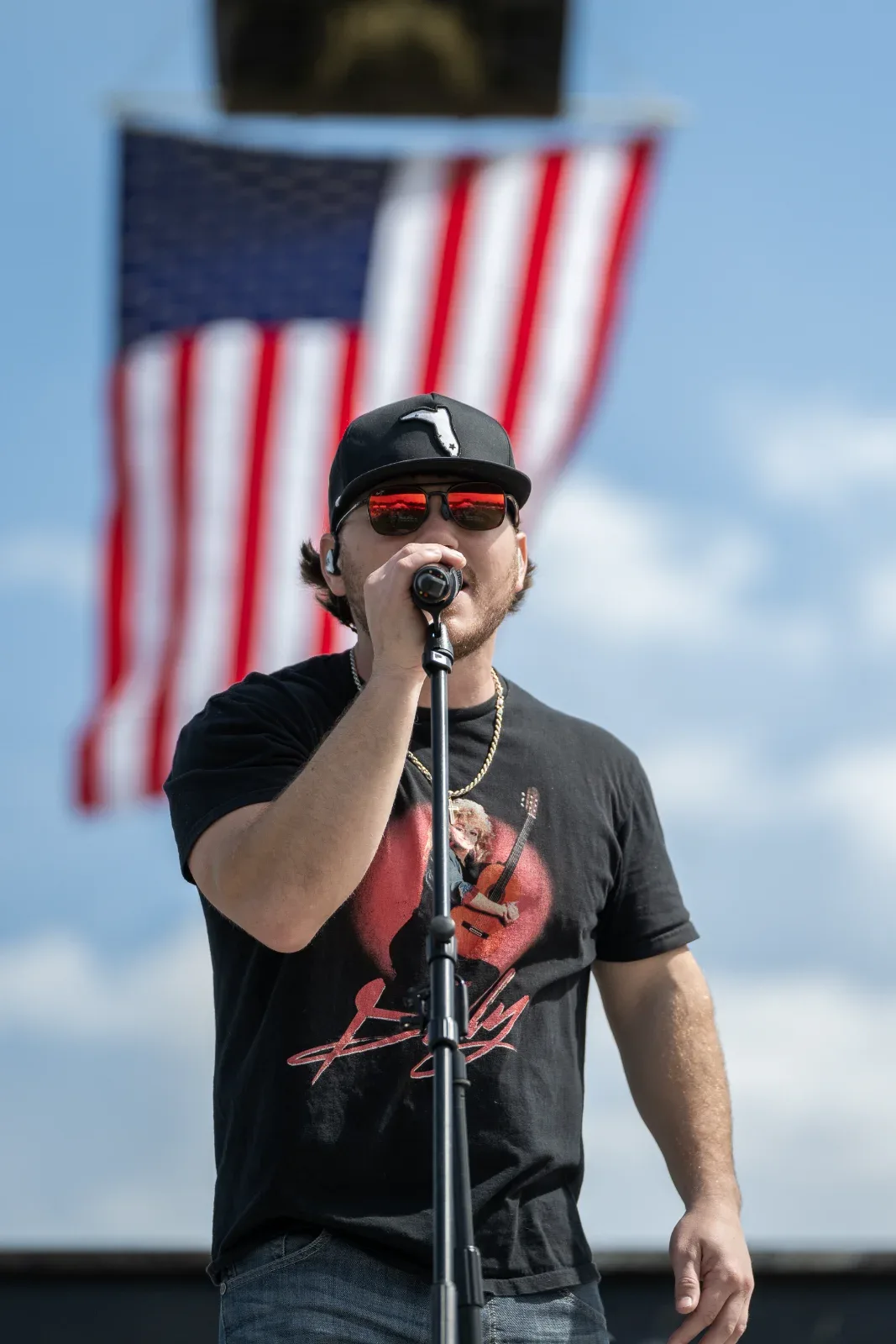A man in a black hat and sunglasses sings into a microphone with an American flag in the background.