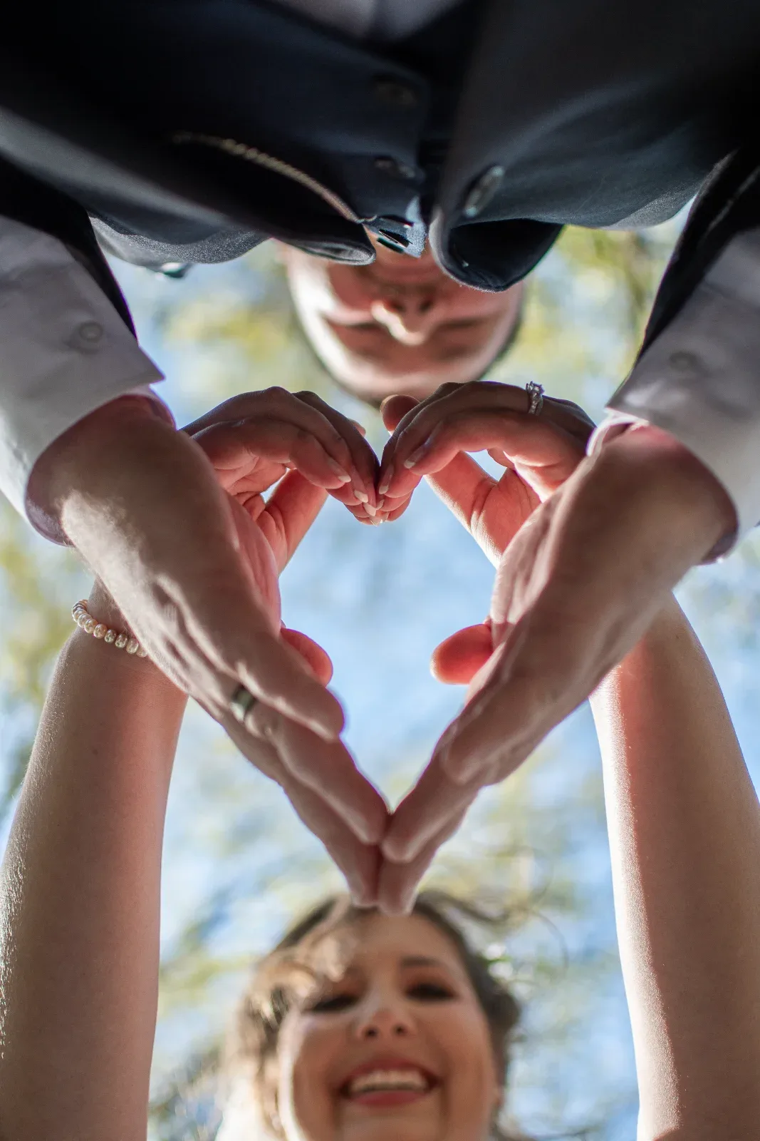 Two people form a heart shape with their hands, seen from below against a blue sky.