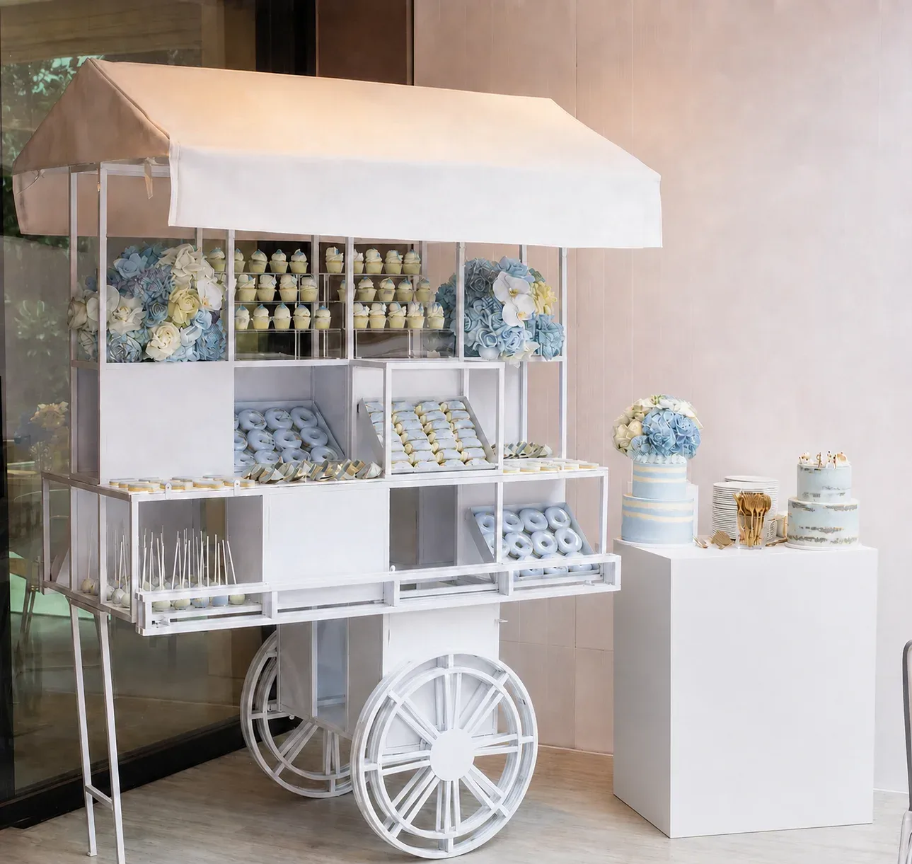 White dessert cart with cupcakes, donuts, and cakes, decorated with blue and white flowers.
