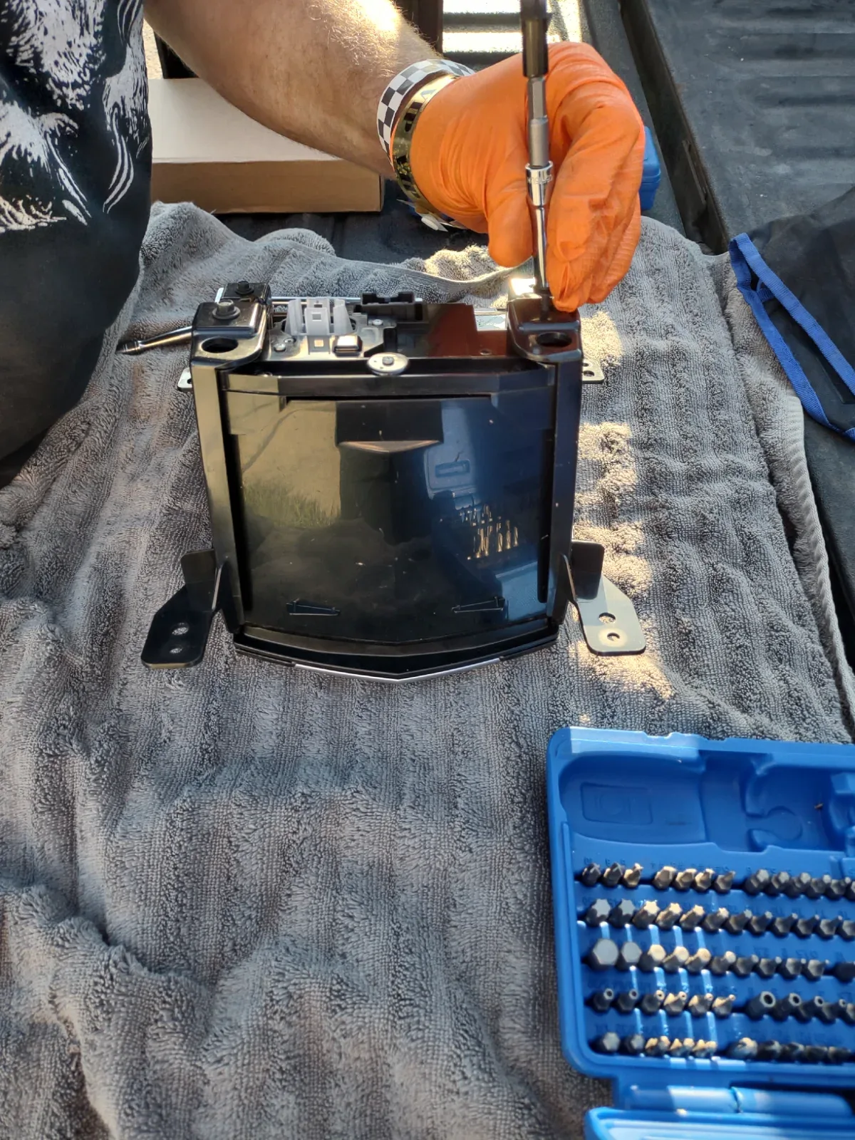 A person in orange gloves uses a screwdriver on a black electronic device resting on a gray towel.