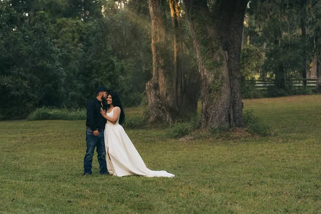 A couple stands in a grassy field with large trees and a wooden fence in the background.