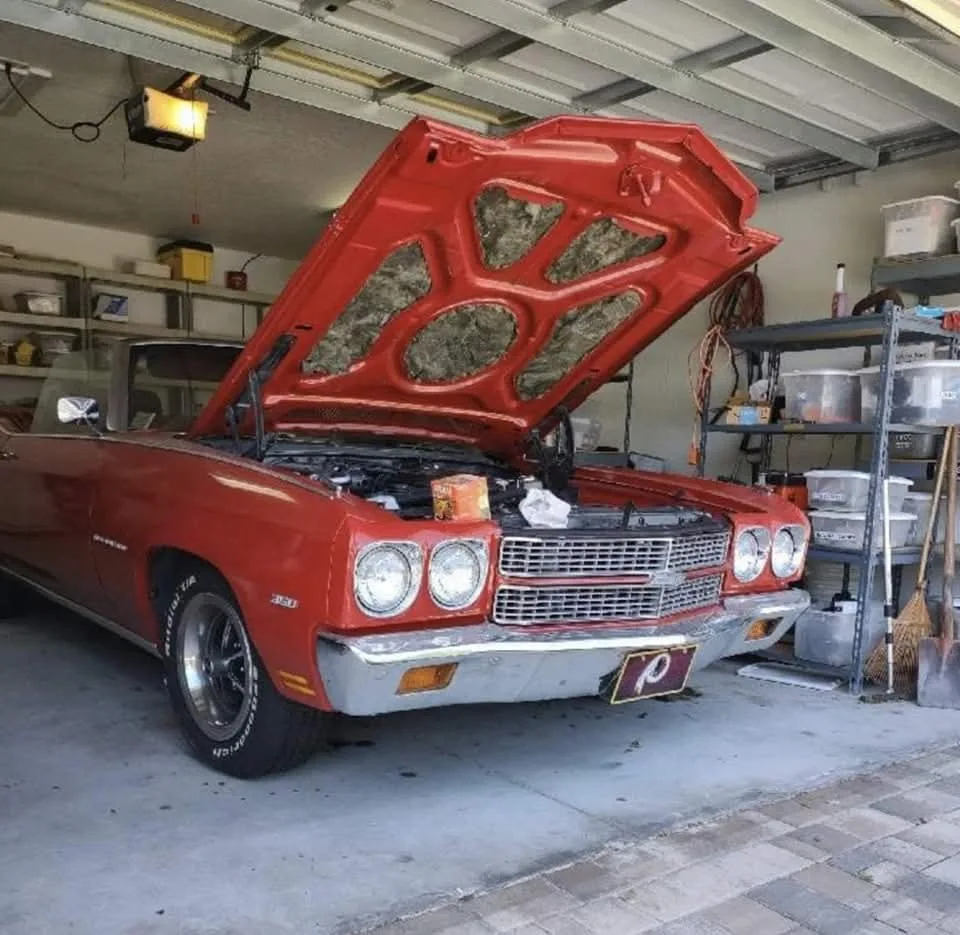 Red convertible car with its hood open in a garage with shelves of items.