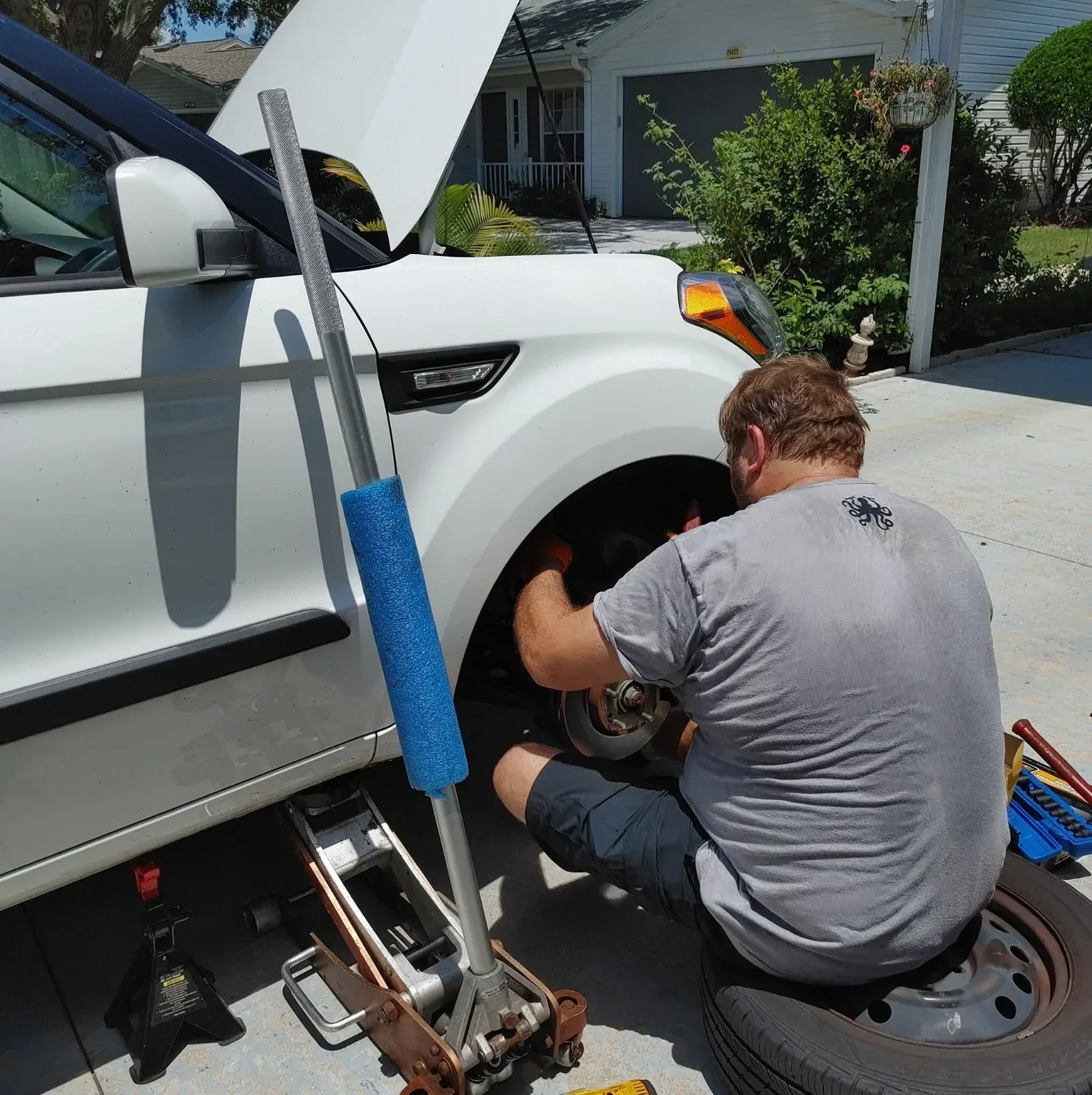 A man works on the front wheel of a white car with its hood open, in a driveway.