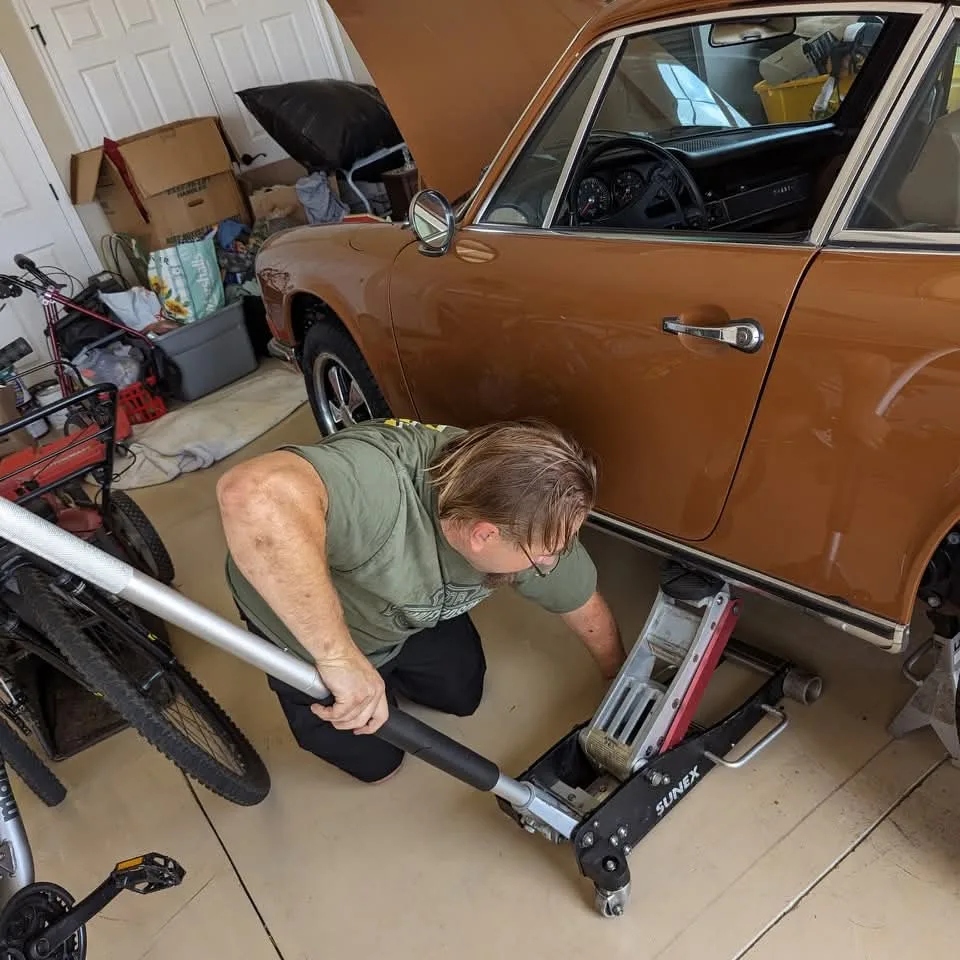 A person kneels, operating a floor jack under a brown car with the hood open in a garage.