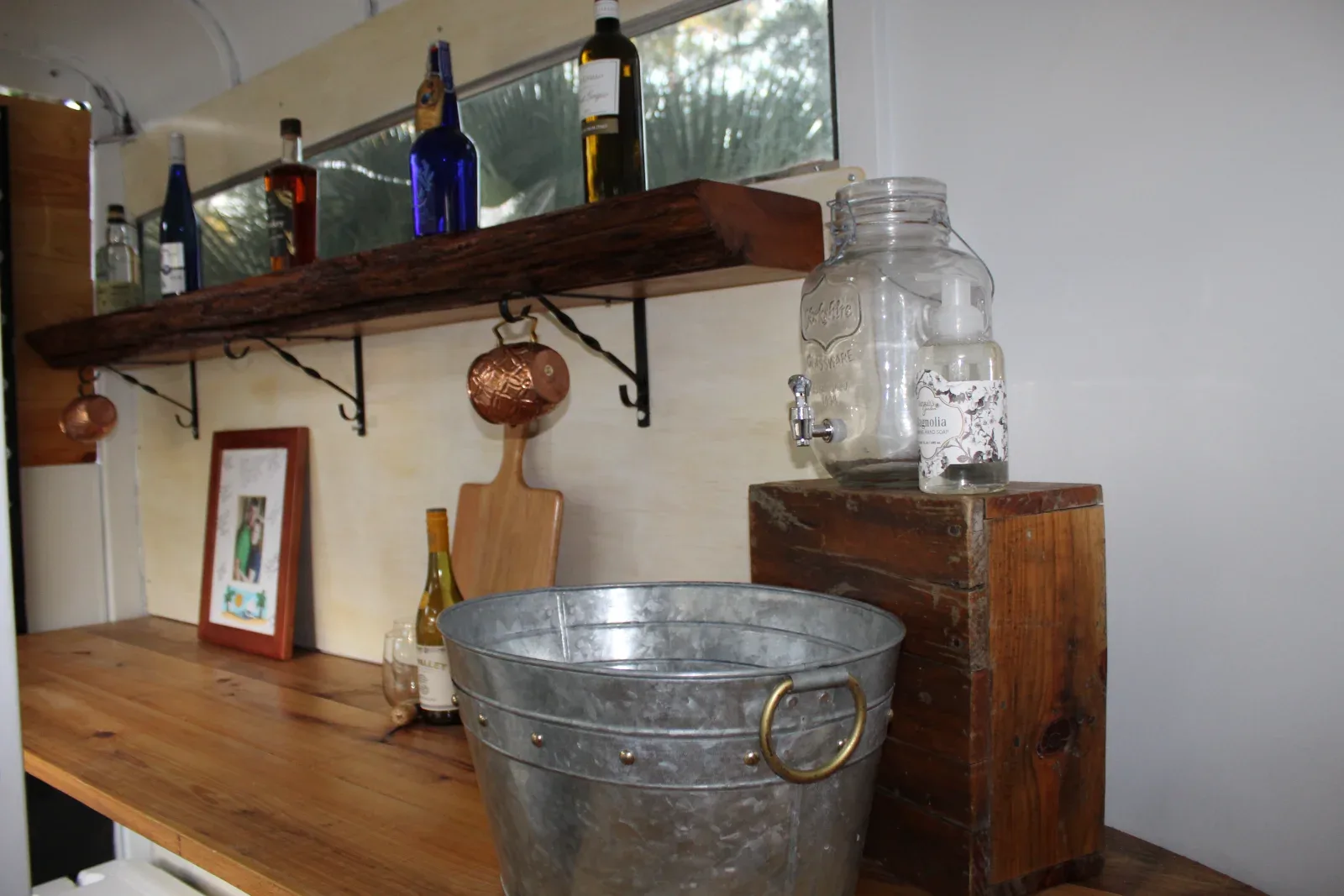 A wooden counter with a metal bucket, a water dispenser, bottles, and a shelf with more bottles.