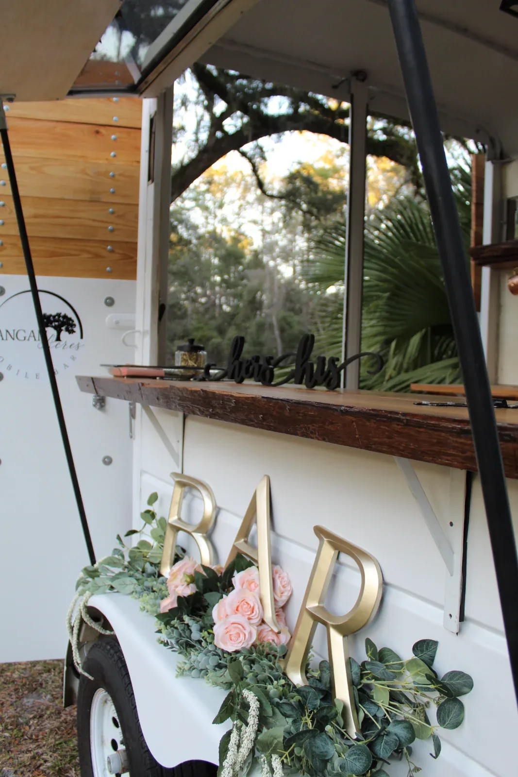 A white mobile bar with a wooden counter, decorated with gold "BAR" letters and pink roses.