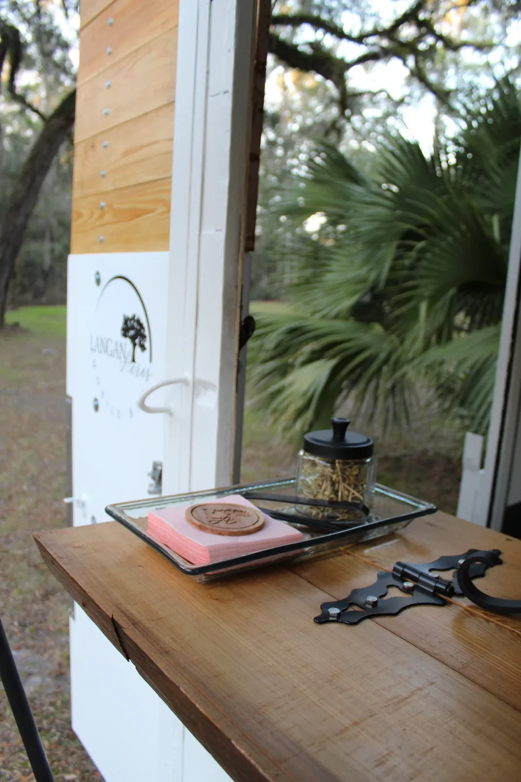 A wooden counter with a glass tray holding pink napkins and a jar of straw, next to a white wall with a logo.