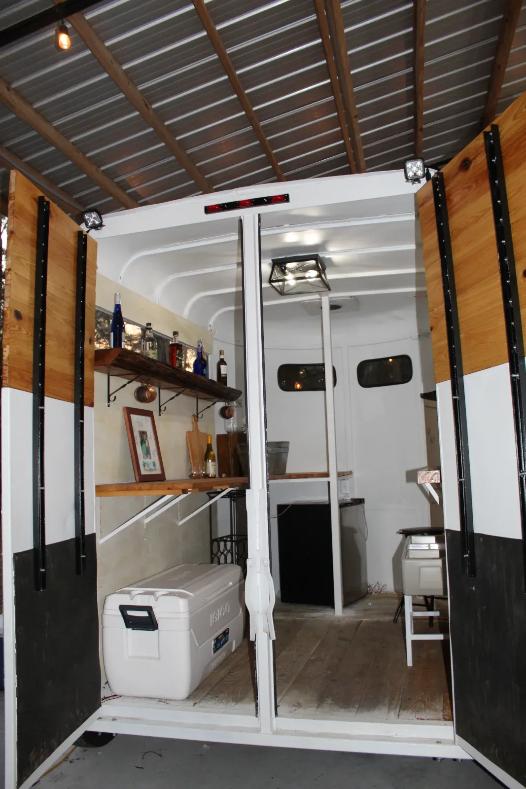 An open white trailer with wooden shelves, bottles, and a cooler inside, under a metal roof.