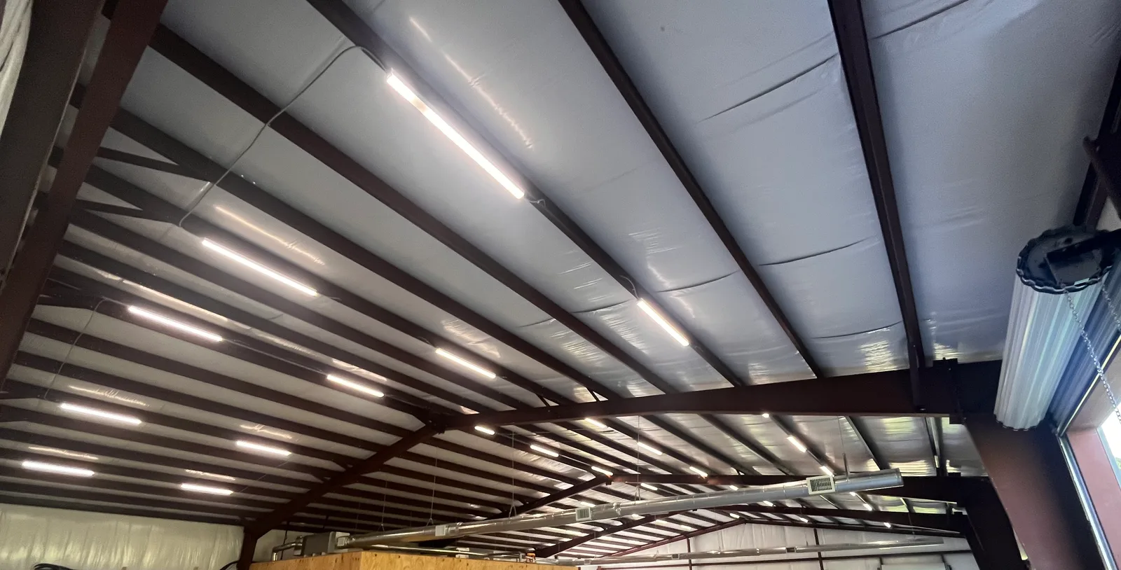 The interior ceiling of a building with exposed metal beams, insulation, fluorescent lights, and ductwork.