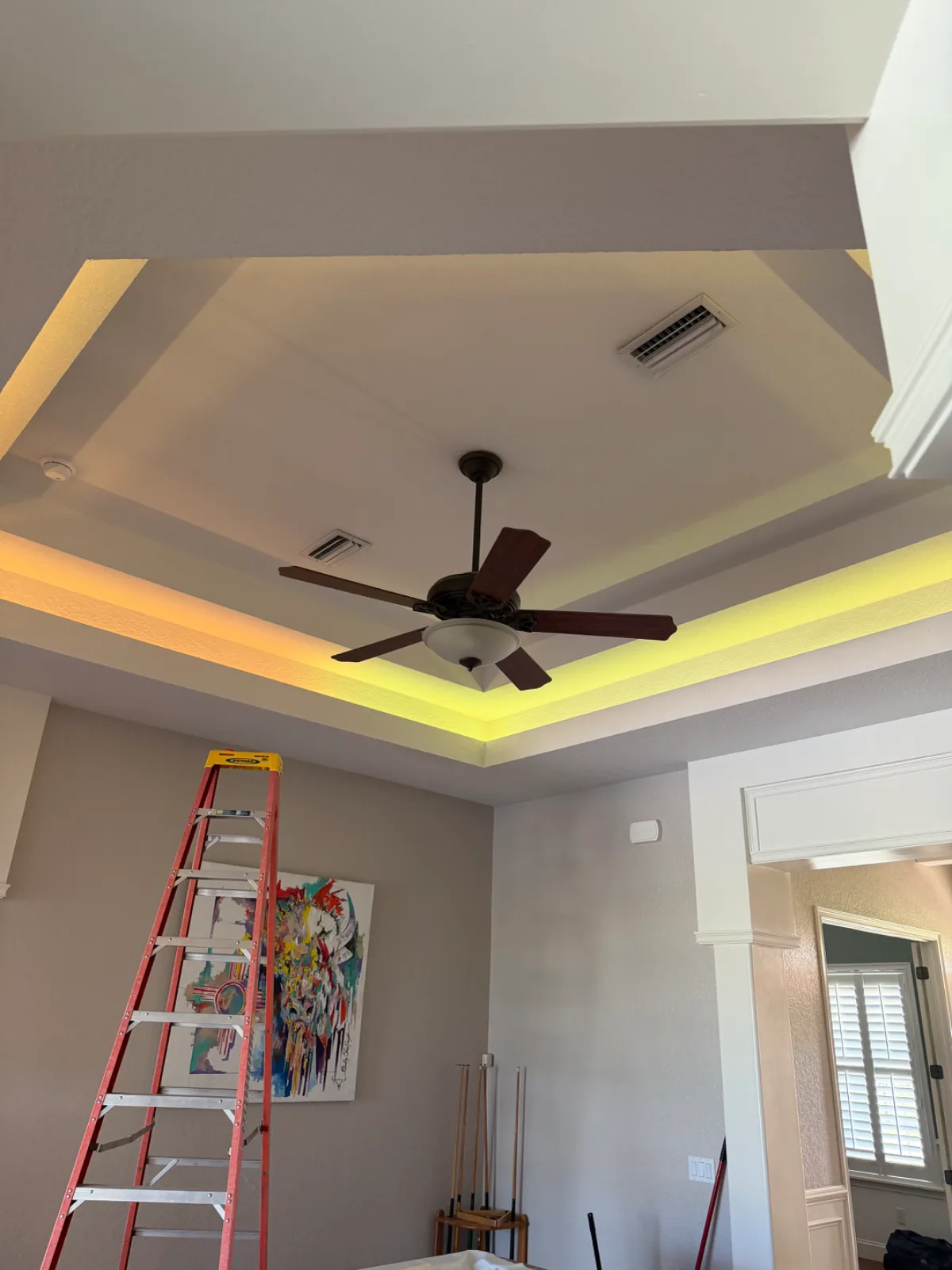 A room with a ceiling fan, yellow cove lighting, a red ladder, and abstract art on the wall.