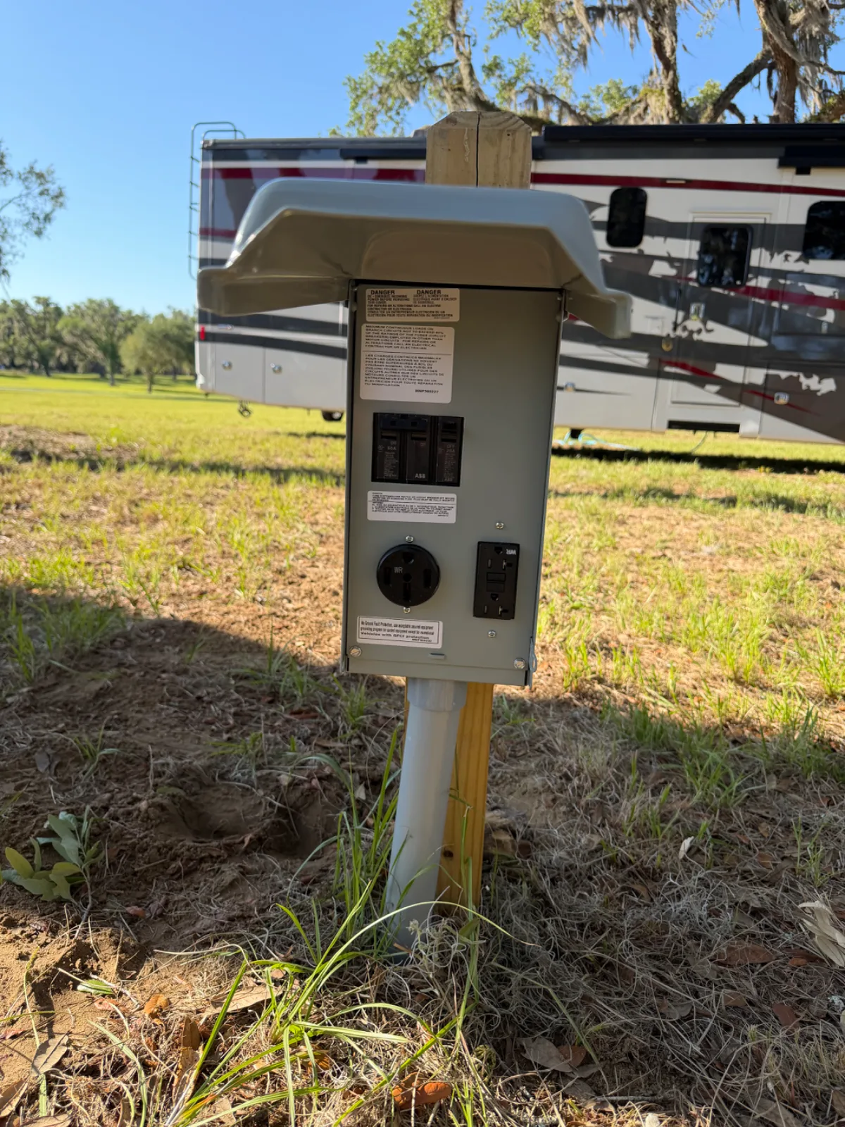An electrical hookup box on a wooden post in a grassy field with an RV in the background.