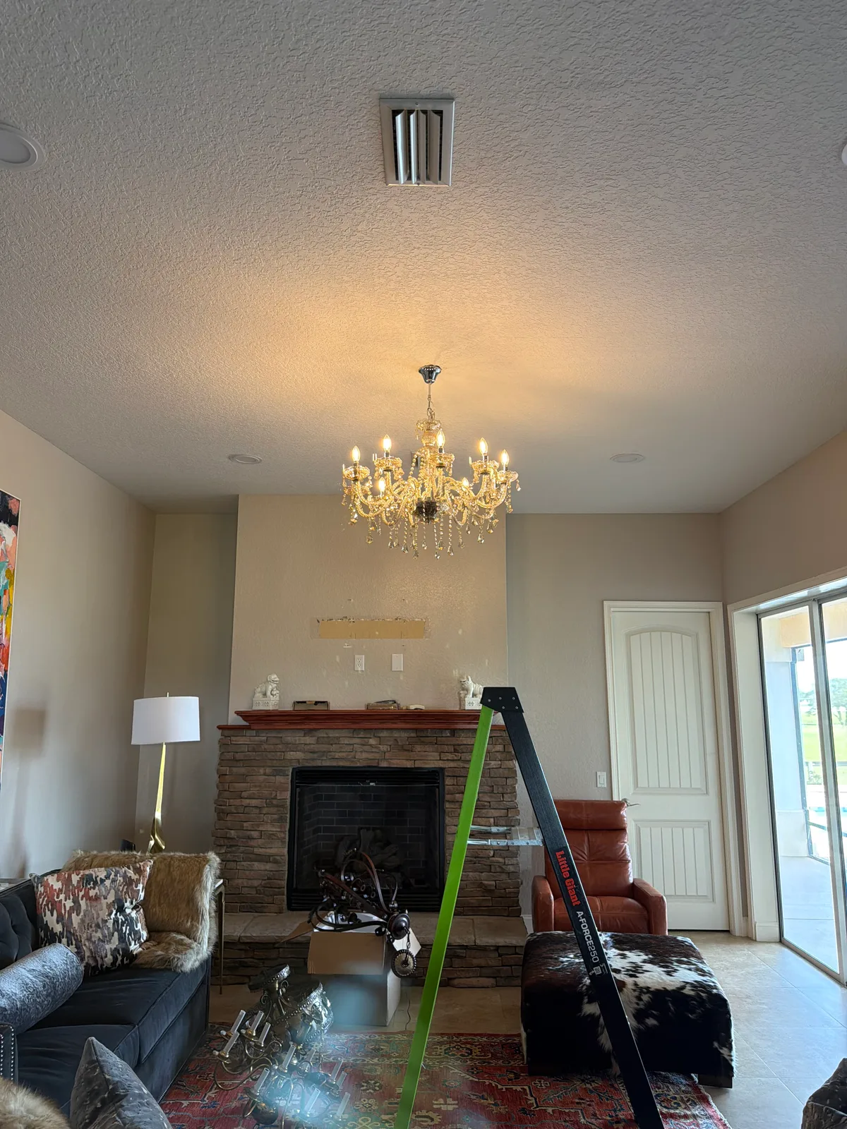 A living room with a lit chandelier, a stone fireplace, and a ladder.