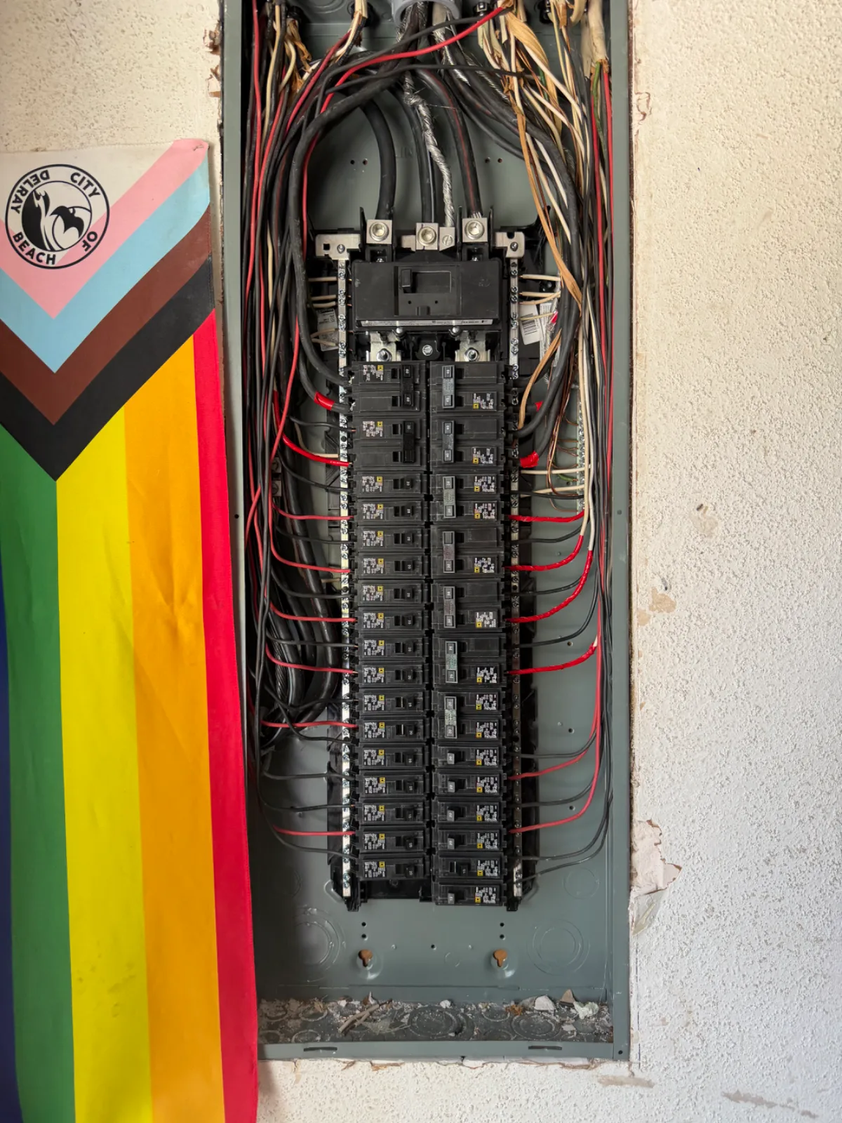 An open electrical panel with many wires and circuit breakers, next to a rainbow flag with a logo.
