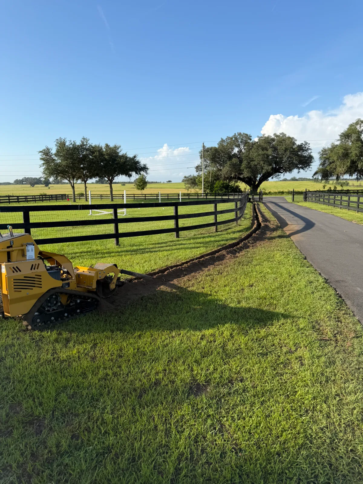A yellow trencher digging a trench along a black fence next to a paved road and green fields.