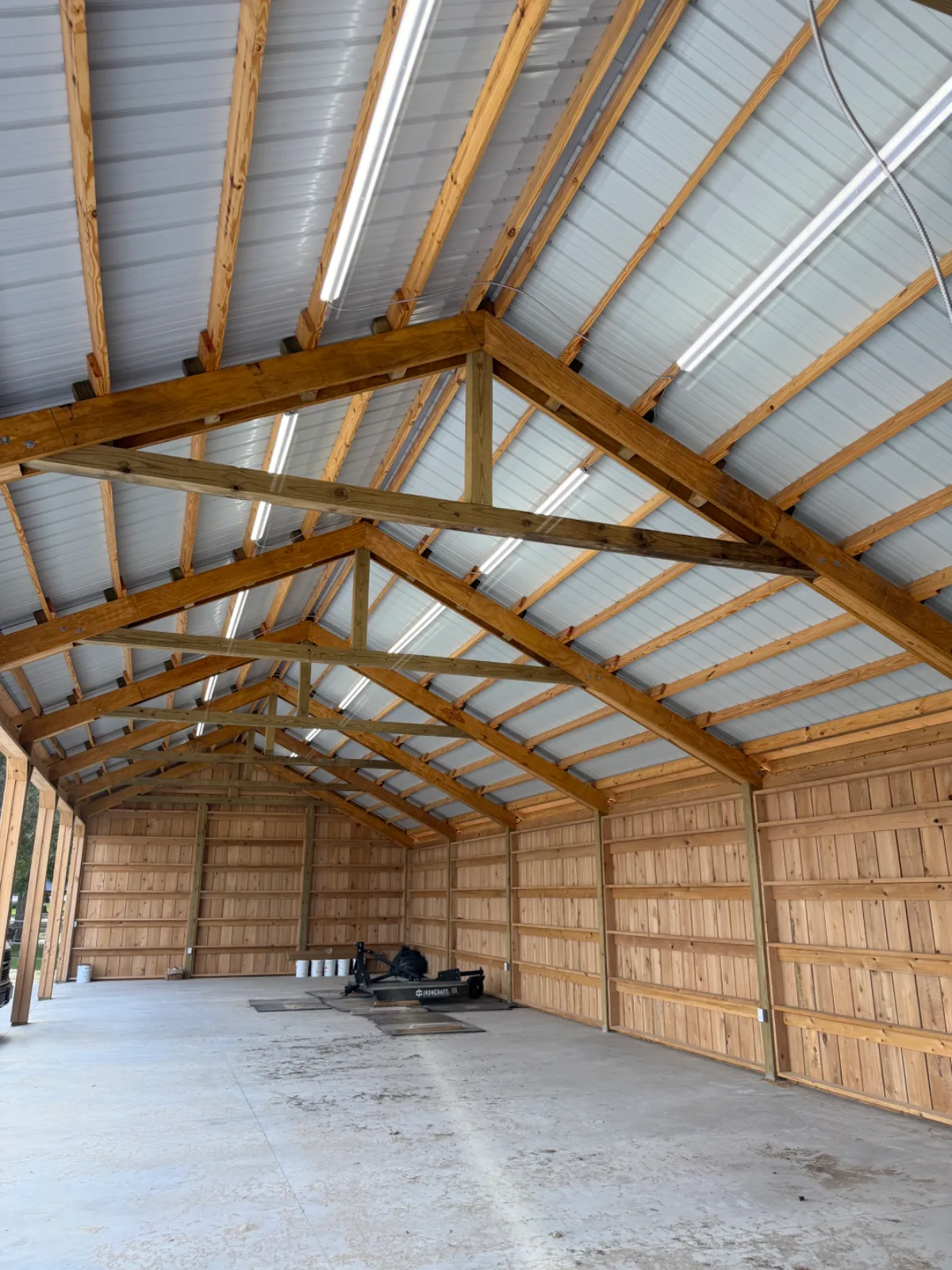 Interior of a large, empty wooden building with a concrete floor and a metal roof with fluorescent lights.
