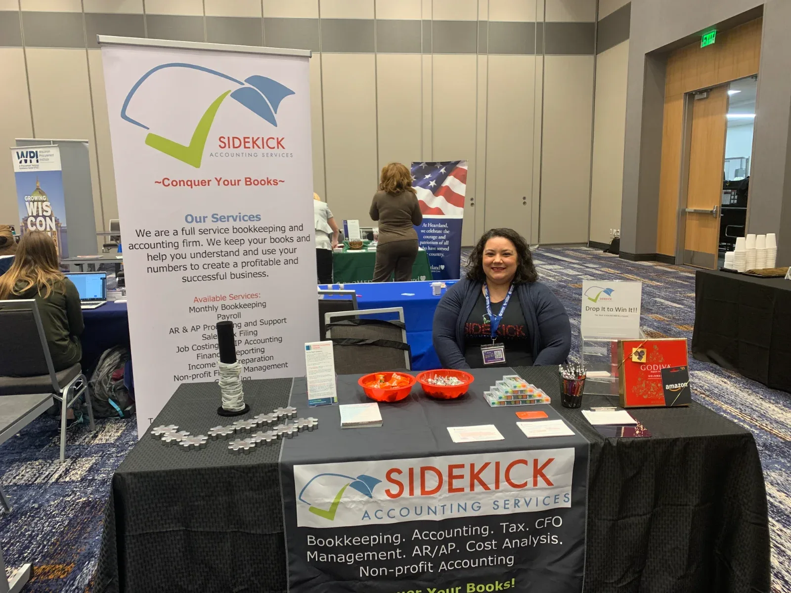 A woman smiles behind a table with "SIDEKICK ACCOUNTING SERVICES" banners and promotional items.