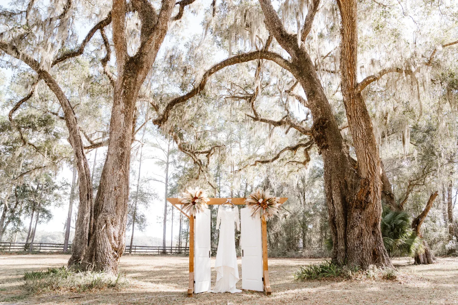 A white wedding dress hangs on a wooden arch decorated with dried flowers, under two large oak trees.