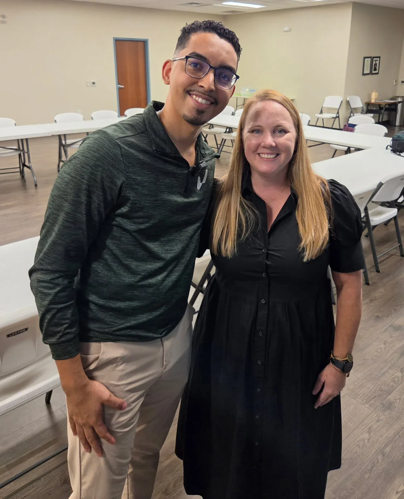 A man in glasses and a woman with long blonde hair smile in a room with white tables and chairs.
