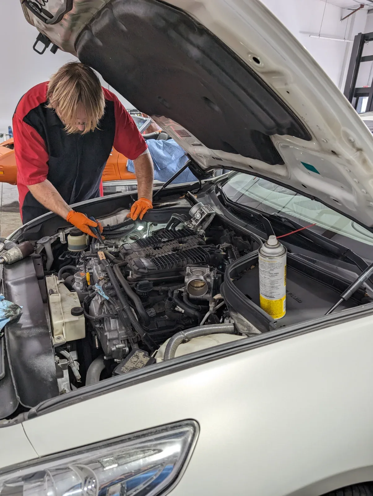 A person in a red and black shirt and orange gloves works on a car engine with the hood open.