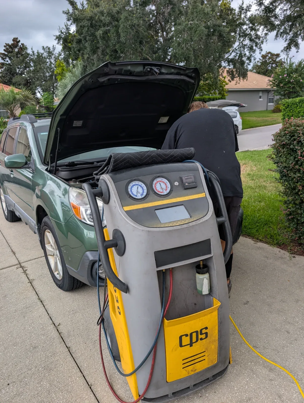 A person works on a green car with its hood open, next to a gray and yellow machine with gauges and "cps" visible.