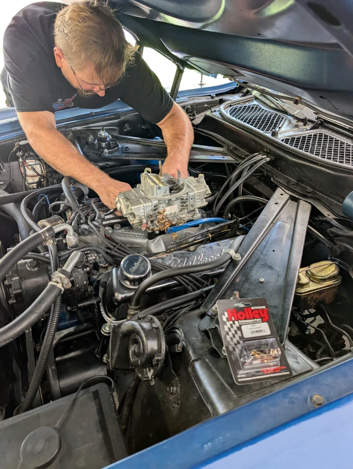 A person works on a car engine with the hood open, a Holley Performance Parts package visible.