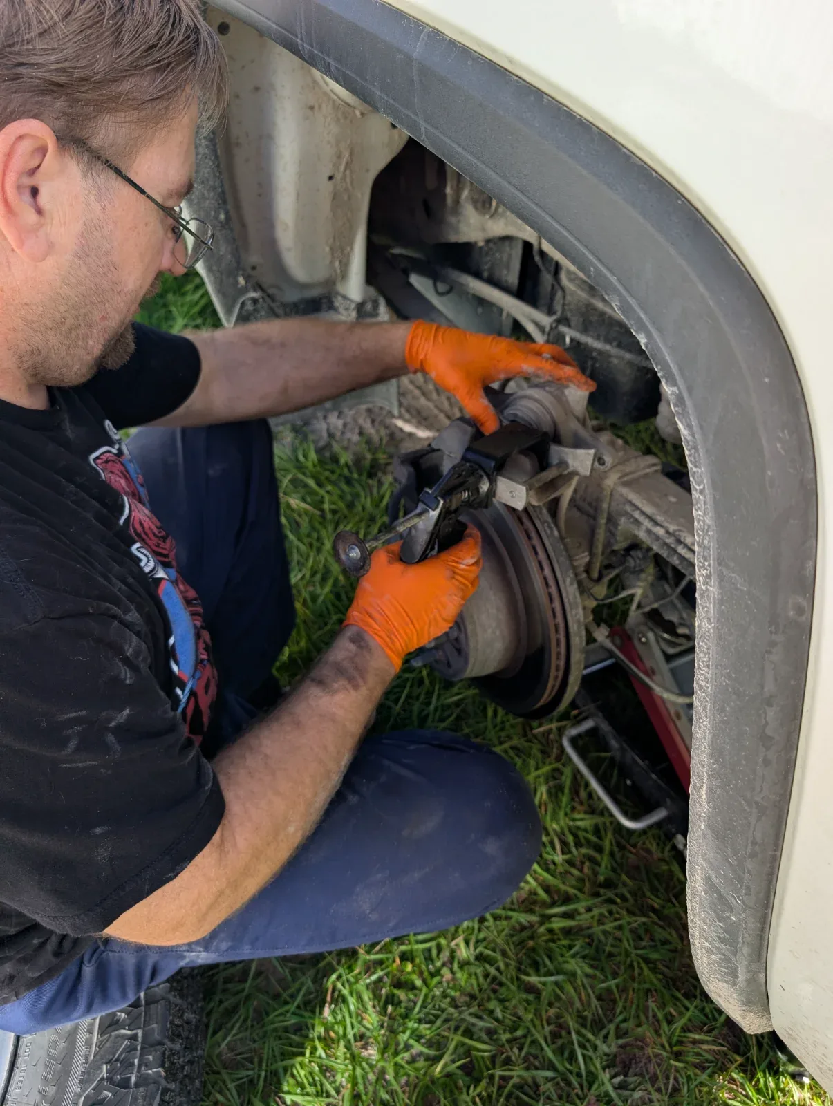 A person in orange gloves works on a vehicle's brake rotor and caliper assembly.
