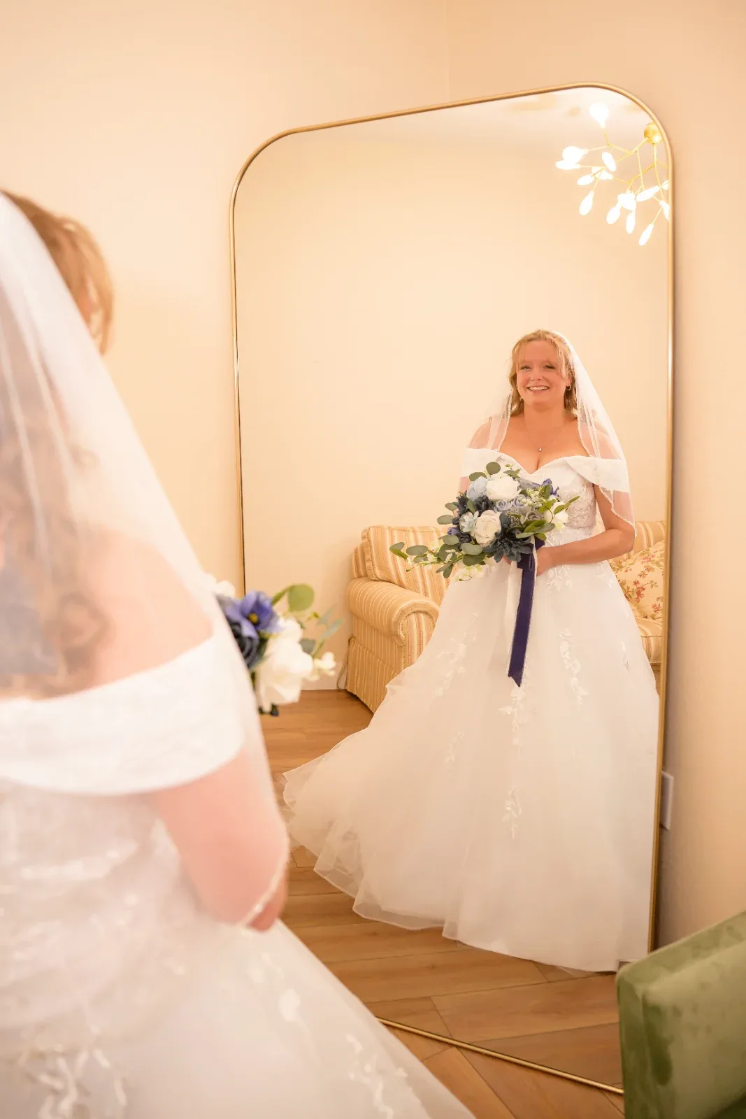 A smiling woman in a white wedding dress and veil holds a bouquet while looking at her reflection in a mirror.