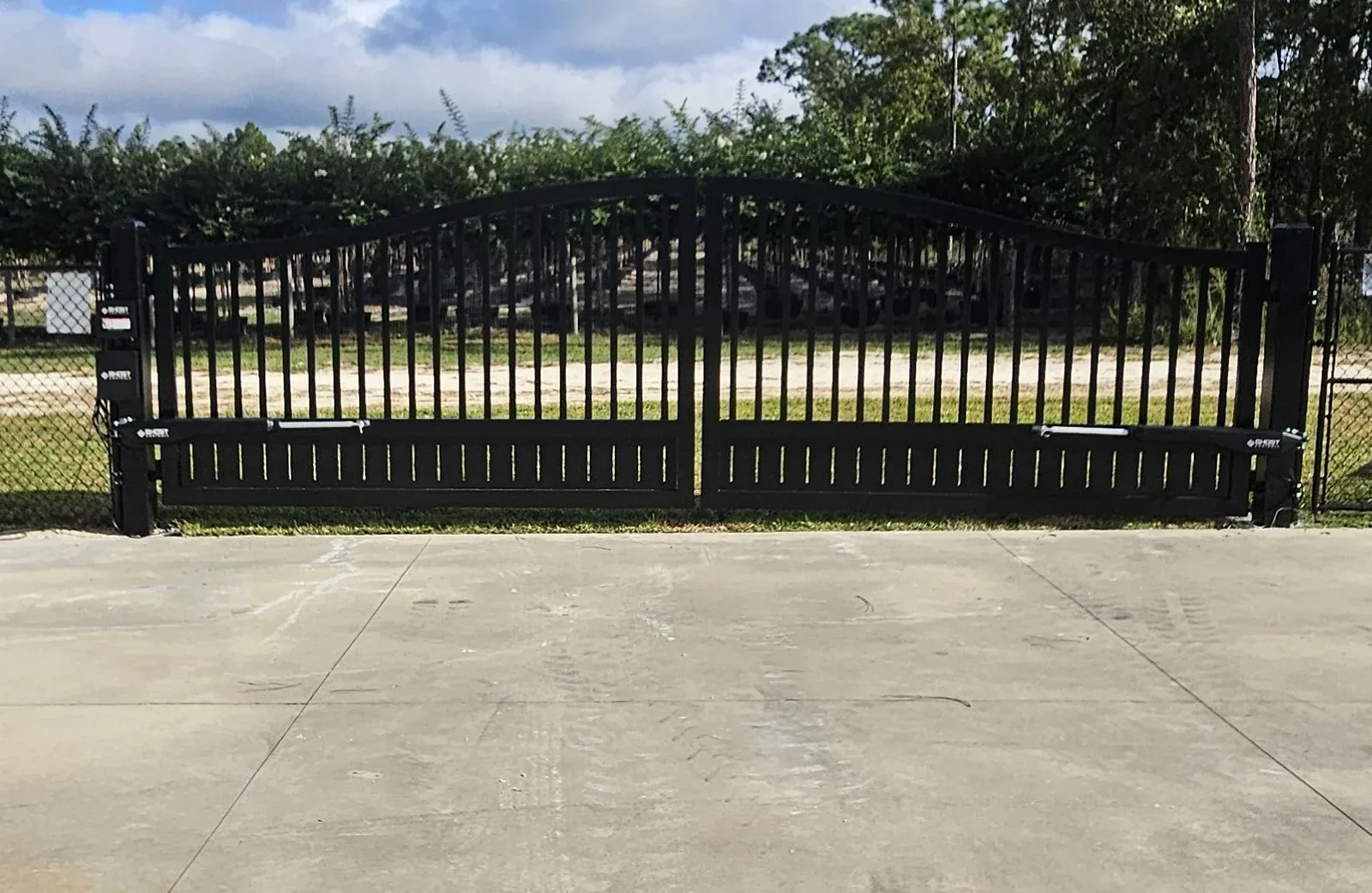 A black metal gate with vertical bars stands closed on a concrete driveway, with green foliage behind it.