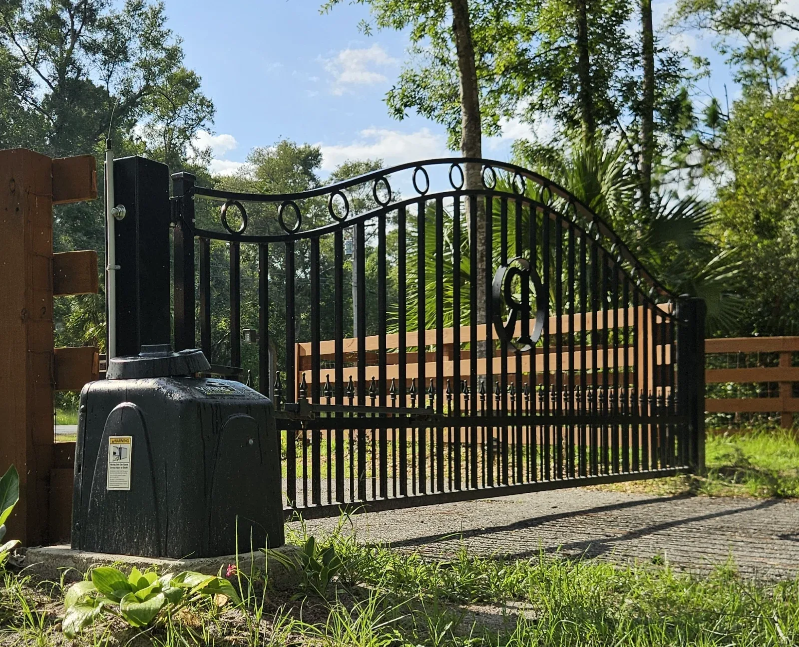 A black metal gate with an automatic opener and a wooden fence, surrounded by trees and grass.