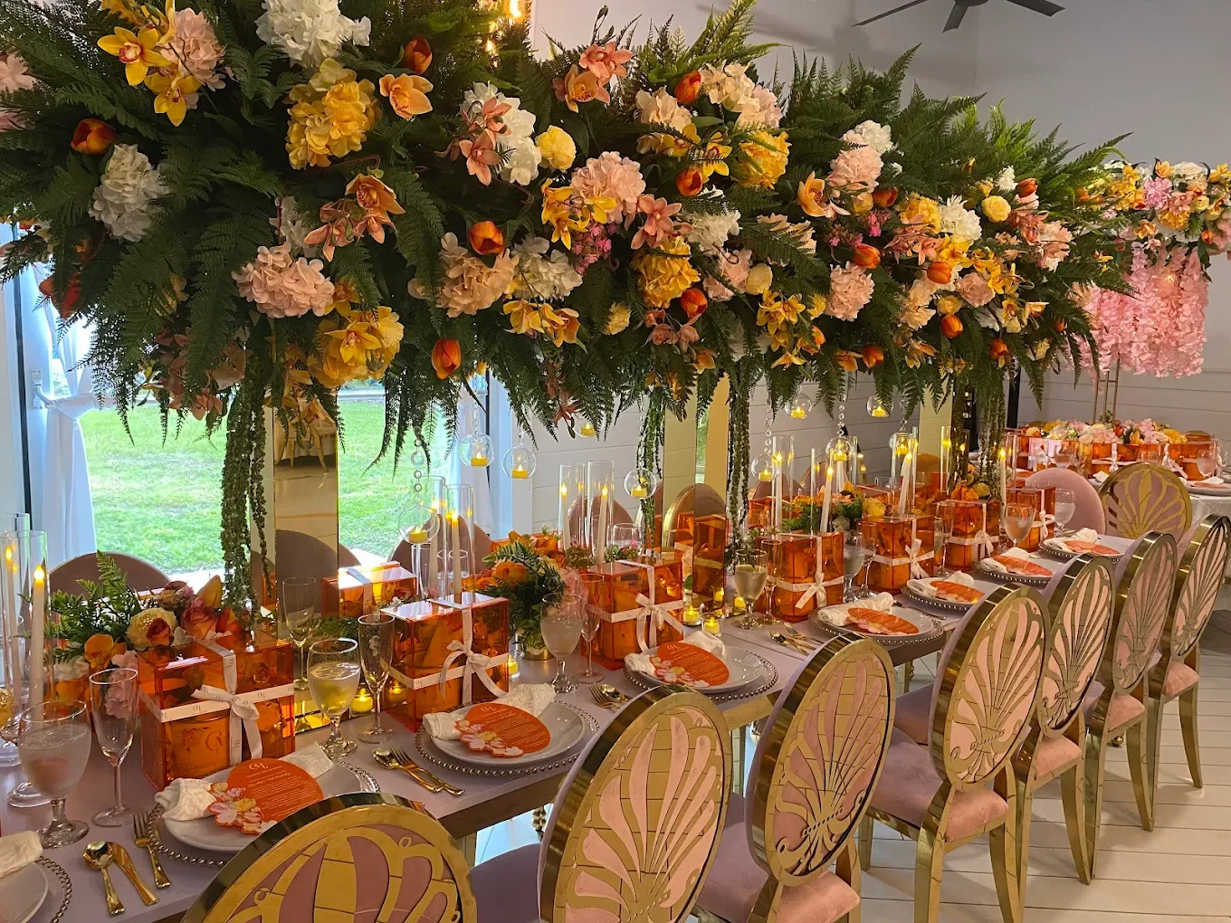 A long table set with orange gifts, candles, and place settings, under a large floral arrangement.